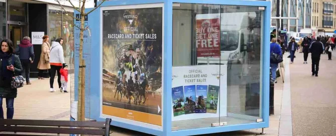 A blue Cheltenham Festival ticket sales pod with horse racing graphics on a paved pedestrianised street.