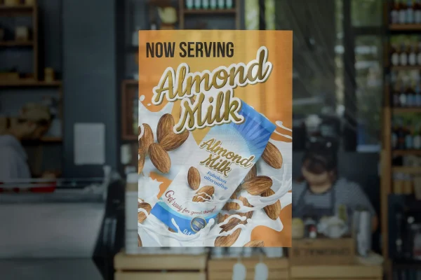 Promotional poster for almond milk featuring whole almonds and a milk splash on an orange background in a cafe setting.