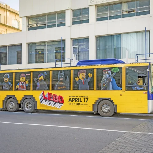 A bus wrapped in bright yellow The Lego Movie advertising with perforated film allowing visibility from inside.