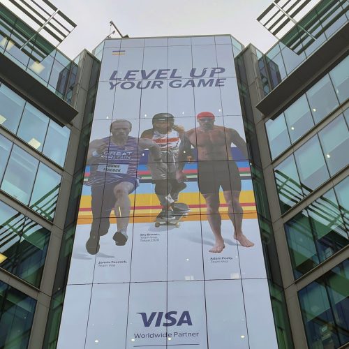 Glass facade featuring Olympian athletes Jonnie Peacock, Sky Brown, and Adam Peaty in a Visa promotional campaign.