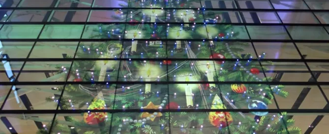 Large glass window panels featuring a brightly lit Christmas tree graphic topped with a crown at Heathrow T2.
