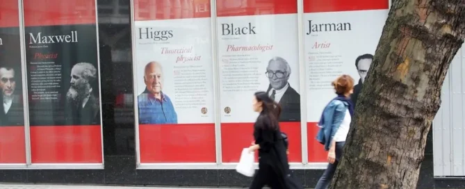 A woman walks past Kings College London exterior windows displaying historical figures like Maxwell and Higgs.