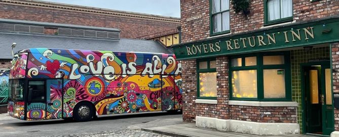 Psychedelic multi-coloured tour bus with 'Love is All' branding, parked by the brick facade of Rovers Return Inn.
