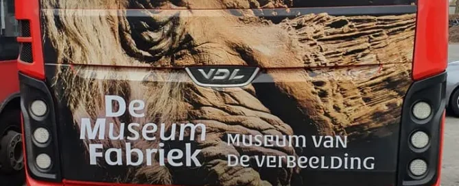 Rear of a red bus featuring a large rhinoceros graphic and De Museumfabriek branding on perforated window film.