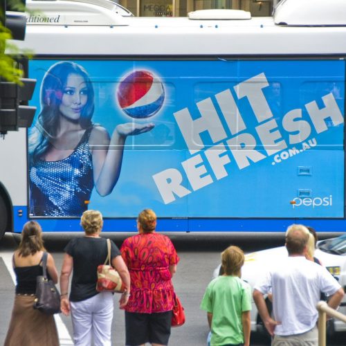 Pedestrians waiting at a junction beside a bus wrapped in a blue Pepsi Hit Refresh advertisement.