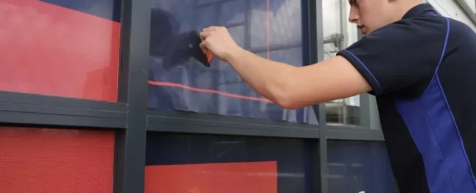 A technician applies red T&A Powering Maintenance window film to an office glass facade.