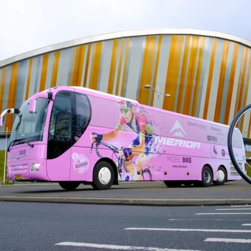 Pink Merida tour bus wrapped in printed film parked outside a modern stadium with a giant pink bicycle.
