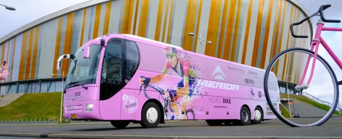 Pink Merida tour bus wrapped in printed film parked outside a modern stadium with a giant pink bicycle.