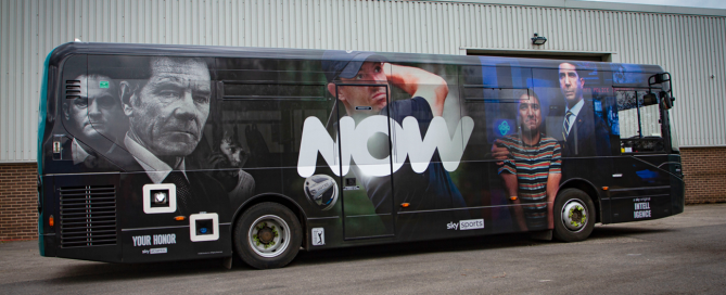 Bus wrap with NOW branding and Sky Sports imagery on perforated window film parked in front of a grey industrial unit.