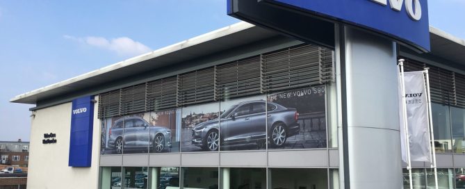 Modern Volvo car dealership with blue signage and large glass windows displaying S90 models, plus cars in the forecourt.