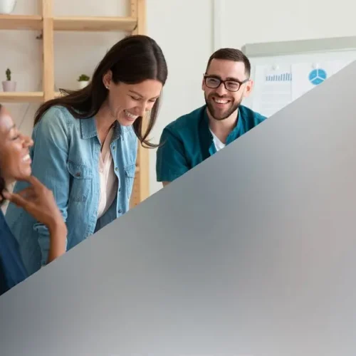 Three colleagues smiling and working together in a bright office behind a translucent frosted glass partition.